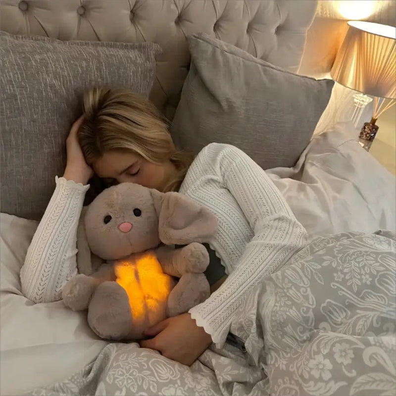 Child lying in bed holding a glowing teddy bear, surrounded by soft bedding and pillows.